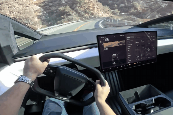 Driver behind the wheel of a Tesla Cybertruck traveling on a winding mountain road with the center touchscreen visible.